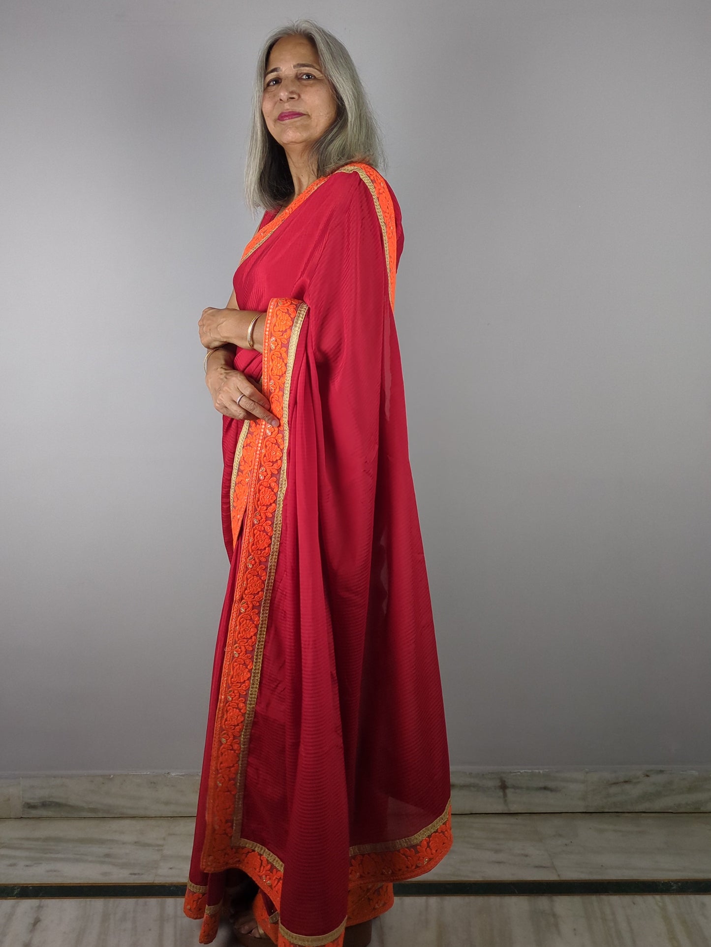 Woman wearing a red saree with orange border against a plain background