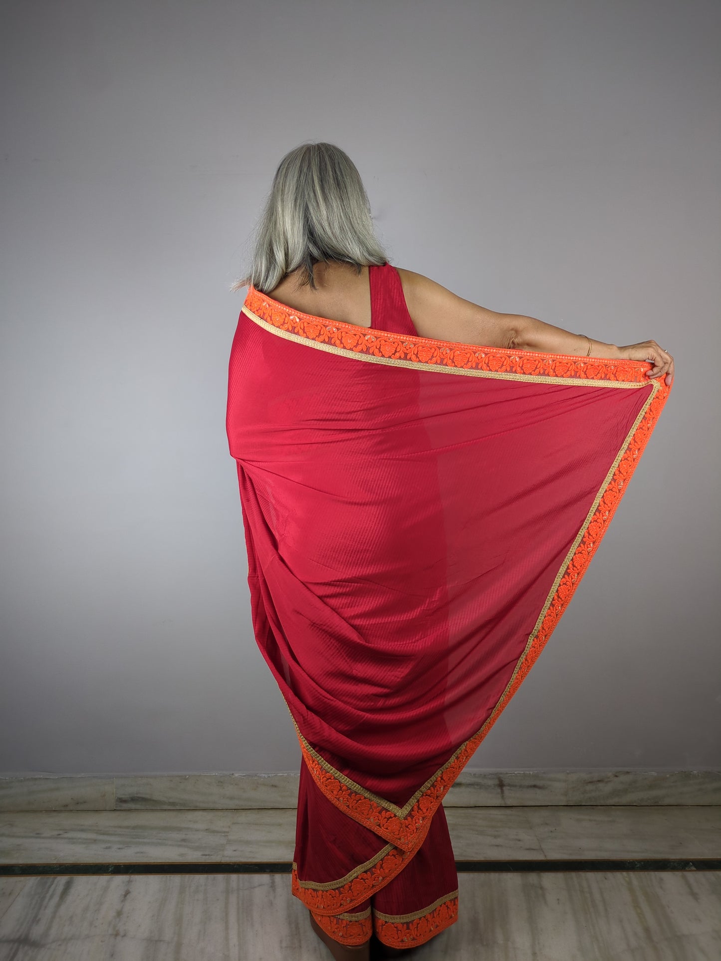 Red saree with orange border draped over a person against a gray background