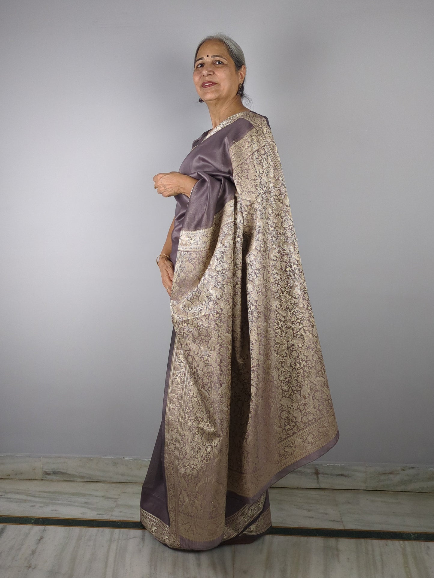 Woman wearing a purple saree with gold border against a plain background