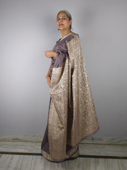 Woman wearing a purple saree with gold border against a plain background