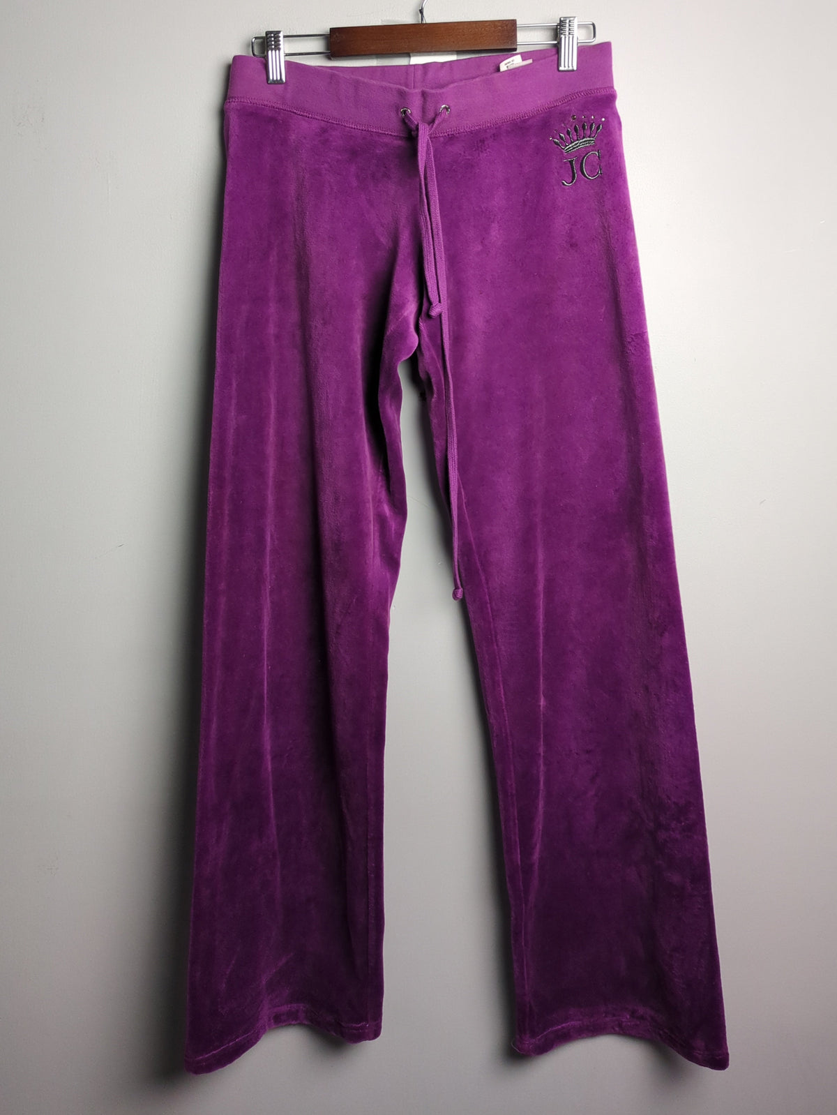 Purple velvet lounge pants hanging on a wooden hanger against a plain light gray wall.