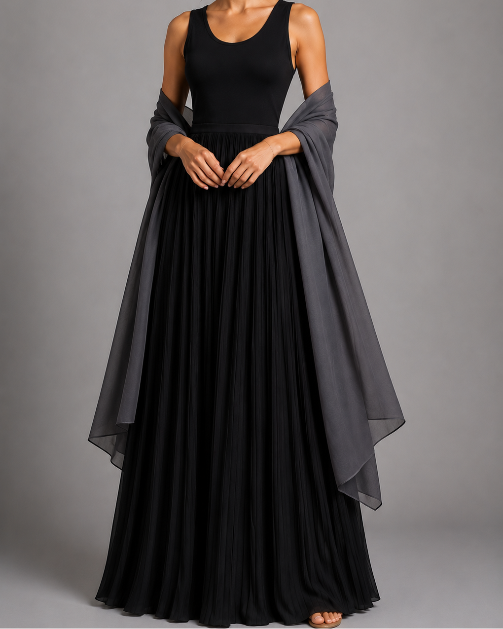 A woman modeling a sleeveless black maxi dress with a sheer dark gray draped overlay, shown against a solid gray studio background.