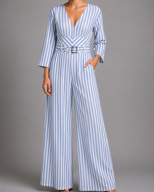 A woman models a blue and white vertically striped wide-leg jumpsuit with a matching belt and silver buckle, set against a solid gray studio background.