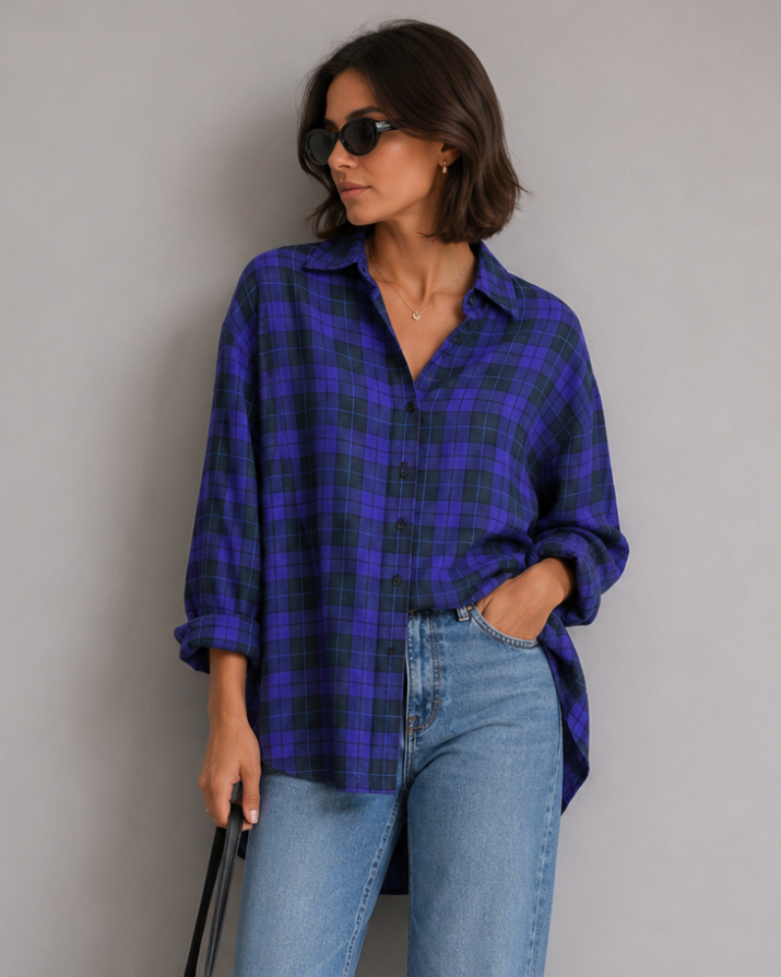 Woman wearing a blue plaid shirt and jeans, holding a black bag against a gray background