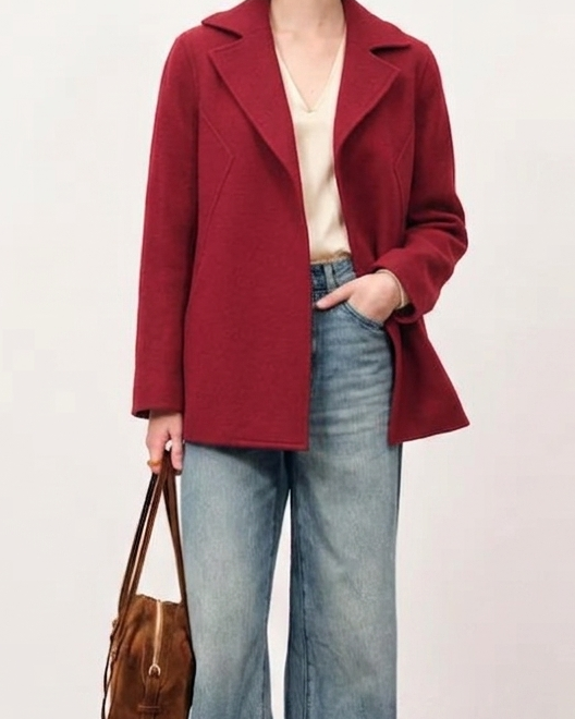 Woman wearing a red coat, light blue jeans, and holding a brown bag on a plain background