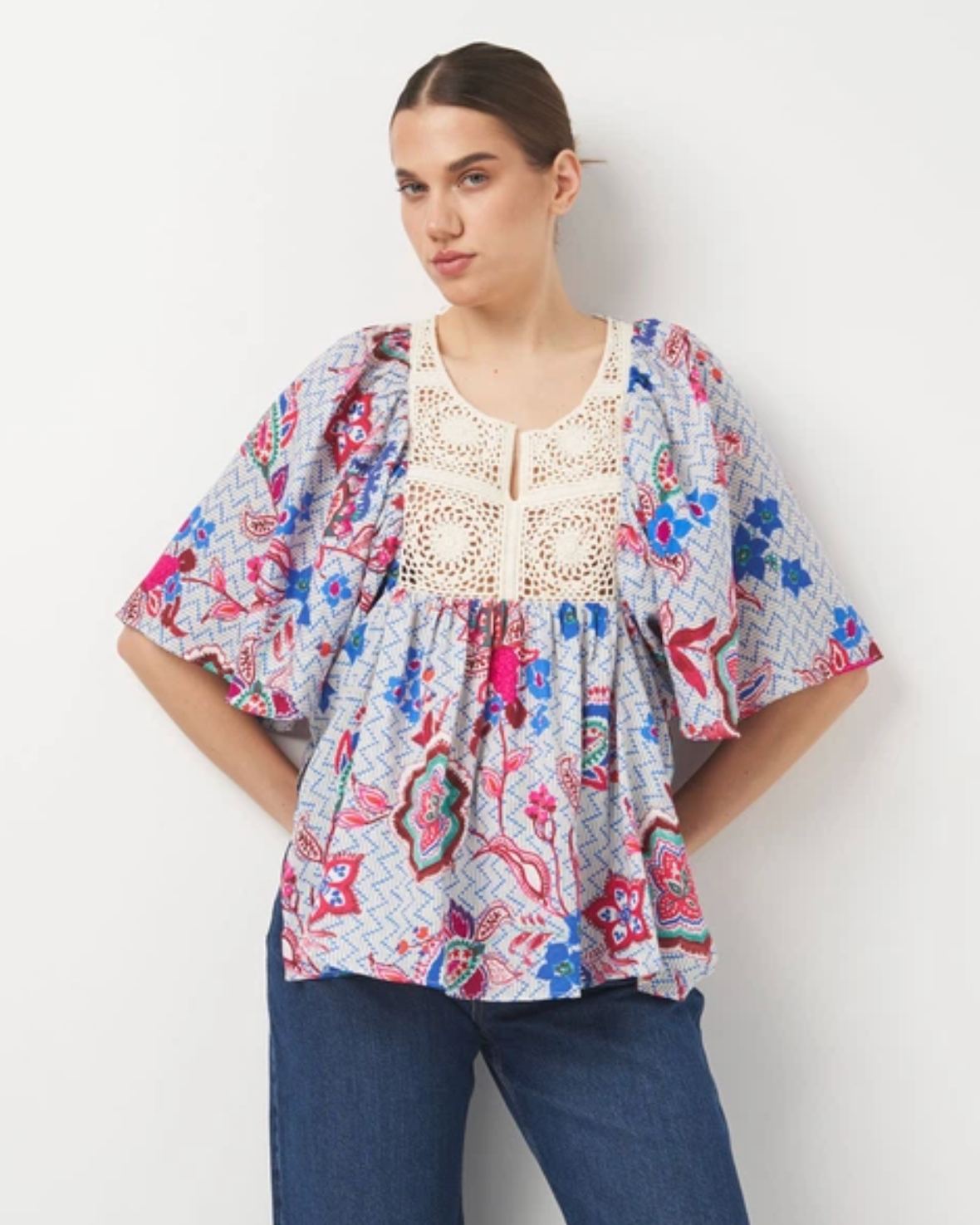 A woman modeling a short-sleeve bohemian-style top with a colorful paisley and floral print on a light blue-gray background, featuring a cream crochet lace neckline. She is wearing blue jeans against a plain white wall.