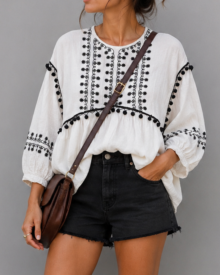 Woman wearing a white blouse with black embroidery and black shorts against a gray background