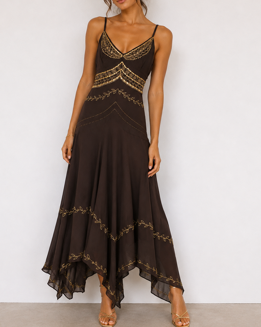 A woman models a dark brown high-low maxi dress with gold embroidered details, paired with gold strappy sandals, against a plain white wall.