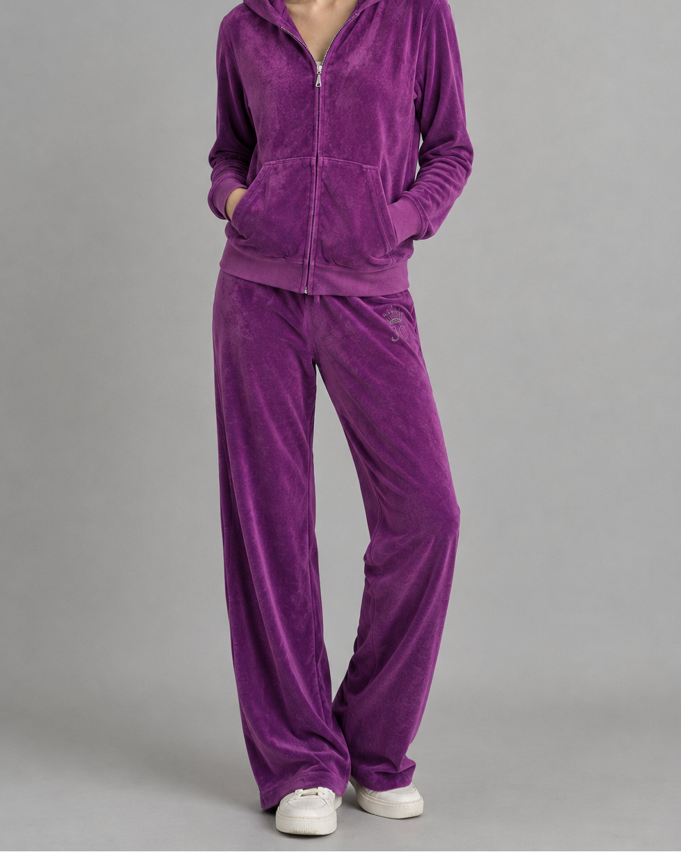 A woman modeling a matching purple velour tracksuit with a zip-up hoodie and wide-leg pants, paired with white sneakers, against a solid gray studio background.