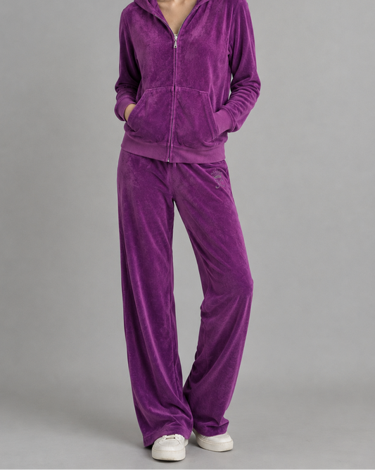 A woman modeling a matching purple velour tracksuit with a zip-up hoodie and wide-leg pants, paired with white sneakers, against a solid gray studio background.