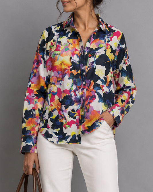Woman wearing a colorful floral blouse and white pants, holding a brown handbag against a gray background