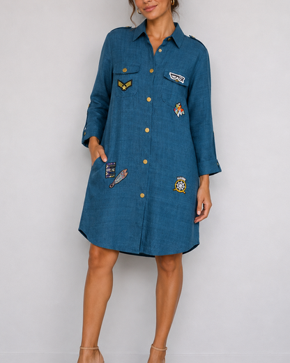 A woman models a blue denim shirt dress with gold buttons and several colorful embroidered patches on the front, paired with beige high-heeled sandals against a plain light gray wall.