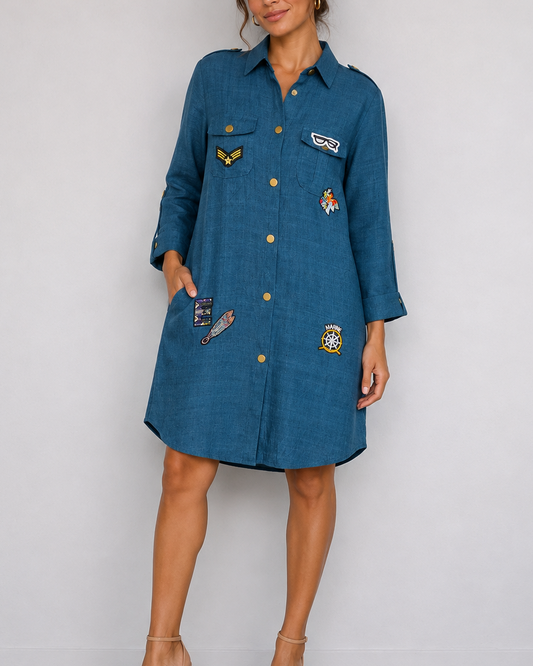 A woman models a blue denim shirt dress with gold buttons and several colorful embroidered patches on the front, paired with beige high-heeled sandals against a plain light gray wall.